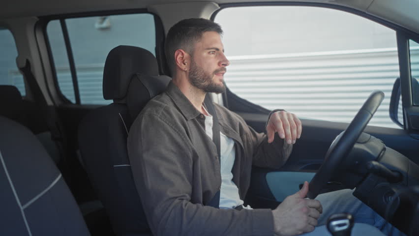 Man rubbing eyes with hand while seated in car next to a building, steering wheel visible; fatigue quiet reflection.