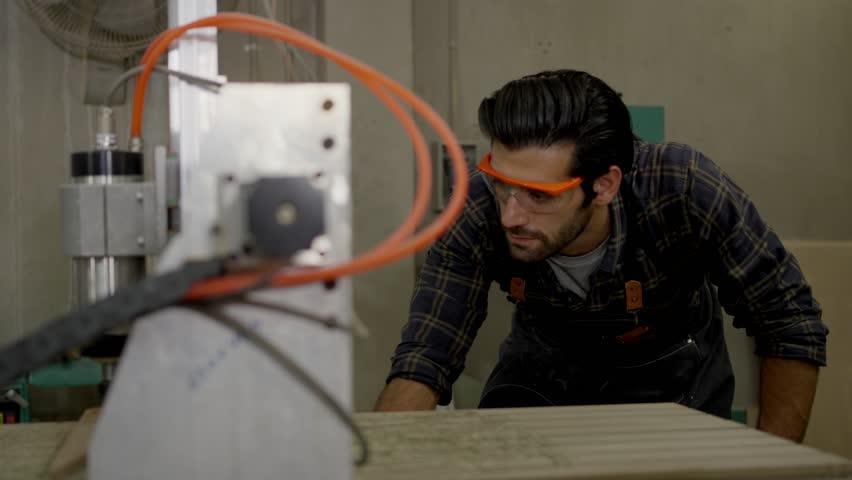 Industrial workshop woodworking scene with safety glasses male carpenter using wood cutting machine and saw on board, inspecting machine setup in factory workshop with industrial equipment and tools