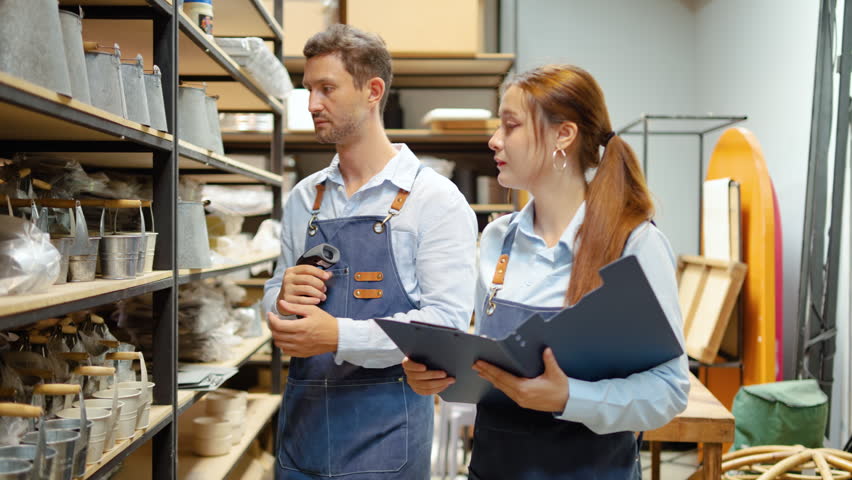 Professional warehouse staff using a barcode scanner to manage inventory of metal pots. Male and female small business owners checking stock levels with a clipboard in a craft store warehouse.