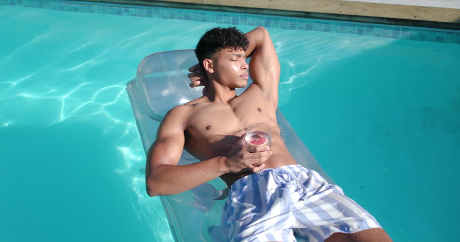 African American man mid-20s lounging on pool float in striped trunks with pink drink shading eyes. Backyard, outdoor, summer, relaxation, serenity, glass, beverage