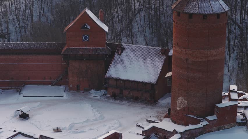 Aerial pass over Turaida Castle in Sigulda, Latvia. A drone sweeps above a frozen courtyard, moving figures by the gate, snow tipped walls, and the frozen Gauja River.