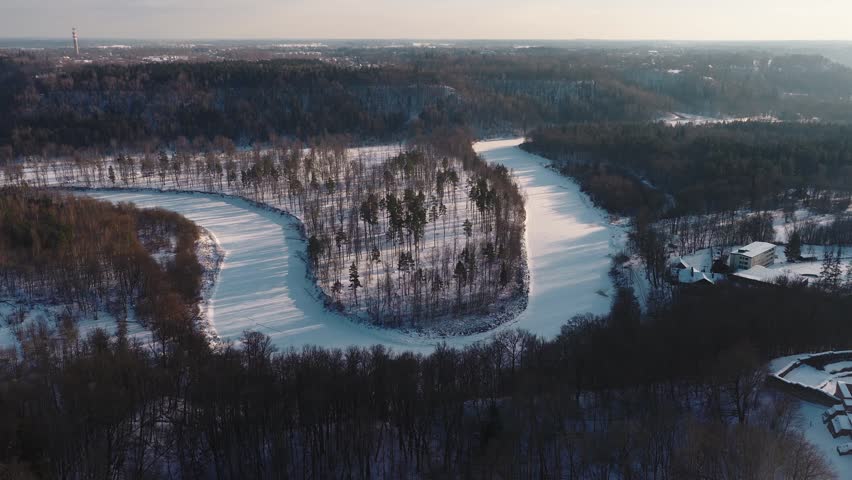 Aerial winter view shows frozen Gauja River curving around a snowy peninsula near Sigulda, Latvia, with Turaida tower on horizon, town, buildings, and long shadows.