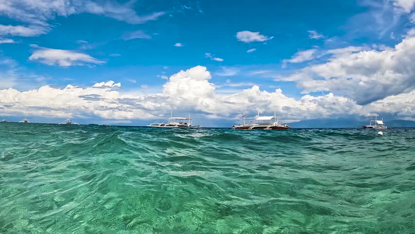 Underwater view of traditional outrigger boats floating on clear turquoise sea water under a cloudy sky in Moalboal, Cebu, Philippines.