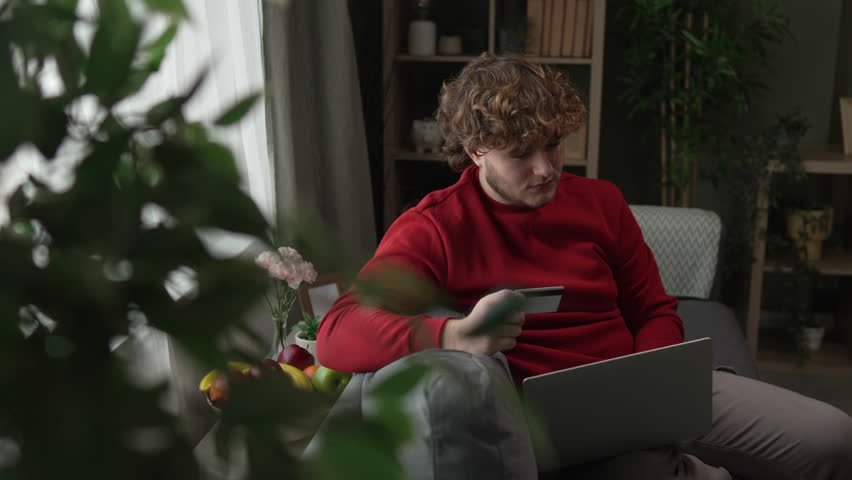 Joyful man with curly hair celebrating a successful online purchase on his laptop, raising his arms in excitement while holding a credit card and sitting on a cozy armchair in his living room