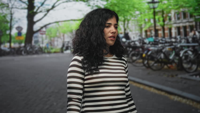 Young woman with hands raised, pained face and striped sweater on street near parked bicycles and trees; frustration resilience.