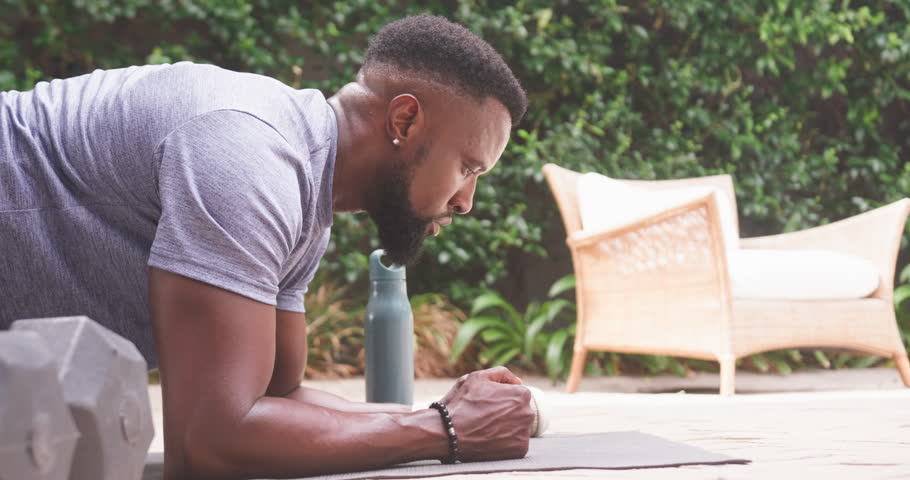 African American man holding forearm plank on mat at patio, wearing grey tee, water bottle nearby. Fitness, outdoor, backyard, focus, balance, strength, dumbbell