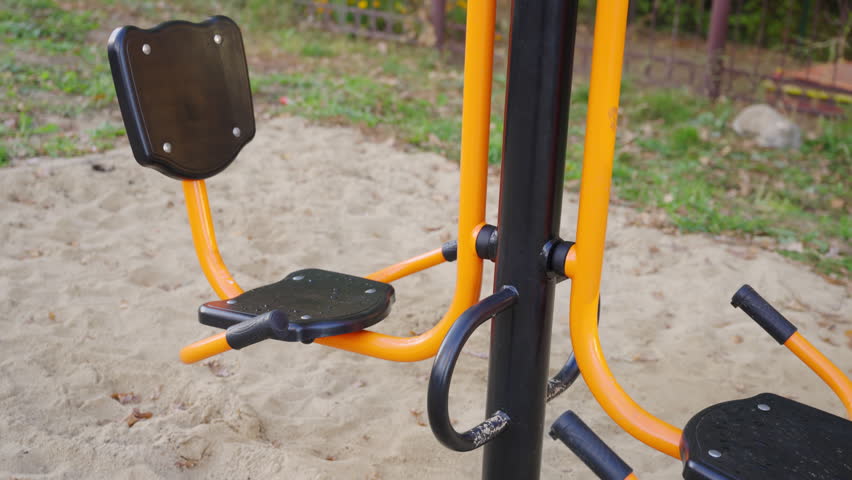 Outdoor fitness seat stands on sand near yellow frame. Exercise station supports seated workouts while offering leg training in yard