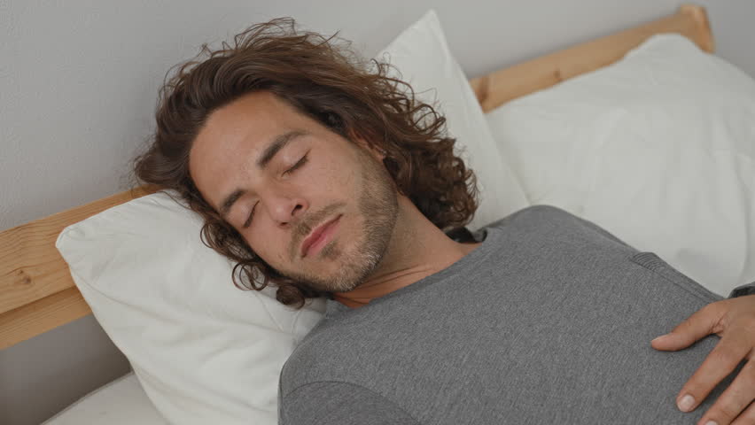 Man reclining on bed with hand on forehead and thoughtful gaze, visible beard and pillow beside him in building; contemplation.