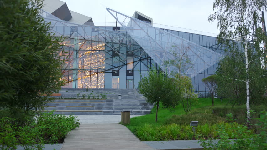 Stylish modern theater building and people group in twilight park. Culture center with illuminated glass facade hosts venue in dusk city