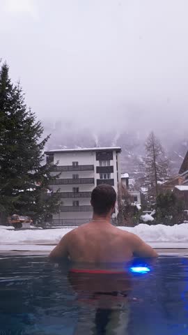 Rear view of a person reclining at an outdoor infinity pool facing a snowy hotel in Zermatt, Switzerland. Falling snow, gentle ripples, fog, and conifer trees appear.