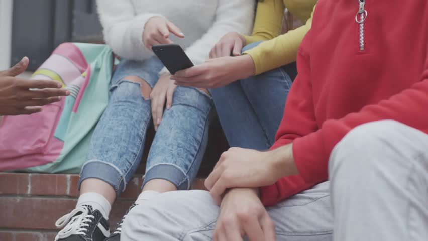 Four teens passing phone on brick steps tapping checking social media metrics appearing over phone. Smartphone, friends, candid, outdoor, campus, daylight, notifications
