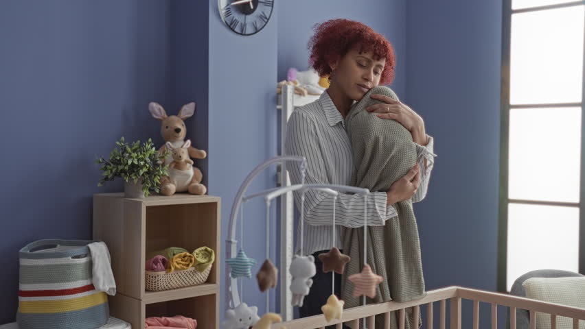 Woman cradling swaddled baby, holding head to chest beside crib and mobile in building nursery; tenderness.