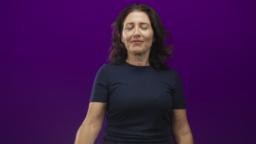 Woman makes l sign with right hand on forehead and sticks out tongue while wearing navy t shirt in studio purple backdrop; playful.