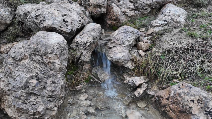 A close-up view of a small, clear stream of water trickling down from between large, textured rocks in a natural, rocky outdoor environment.