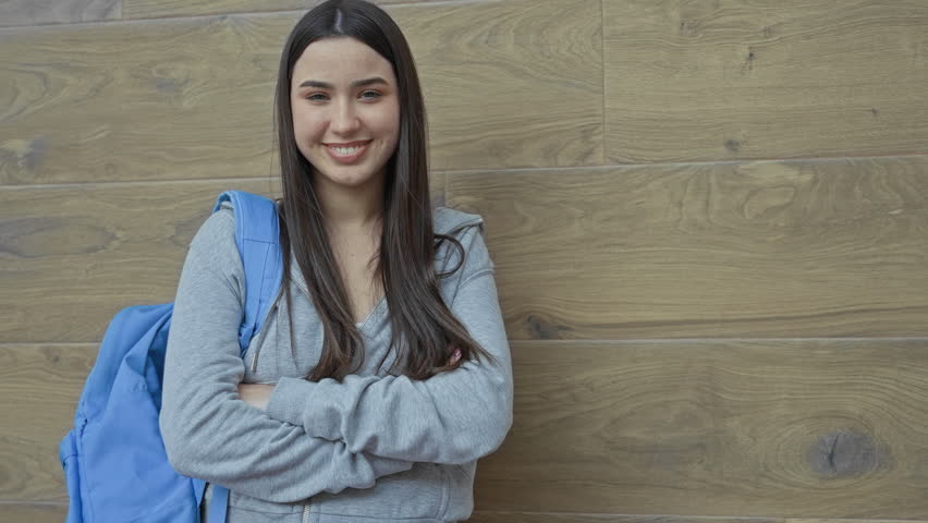 Young hispanic woman leaning against wood wall with blue backpack, arms crossed and smiling in a school building; confidence study optimism.