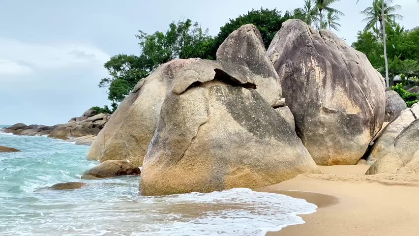 A landscape shot of massive smooth rock formations on a sandy shore, with waves crashing against them under an overcast, rainy sky. Green tropical vegetation and palm trees are visible in the background