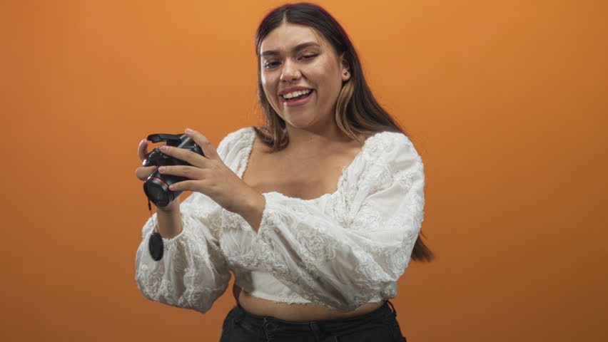 Woman holding dslr camera, hand adjusting lens against orange studio backdrop, smiling and wearing white embroidered blouse; joy creative.