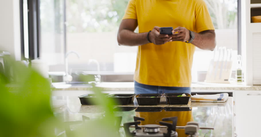 African American man in yellow tee seeing notification, tapping phone at island, sharing four trays. Kitchen, smartphone, counter, mealcontainers, naturallight, plant, scrolling