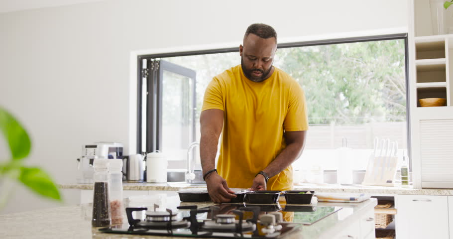 African man leaning picking lids pouring jug sealing black trays prepping food on island near stove. Countertop, mealprep, organized, kitchenware, cooktop, daylight, modern