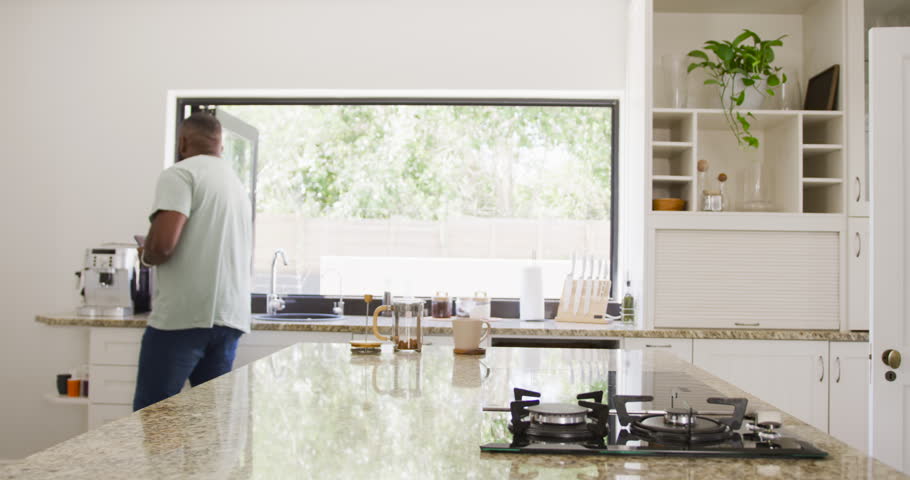 Man starting morning routine in kitchen, pouring hot water into French press while checking phone. Coffee, device, home, modern, natural, light, window