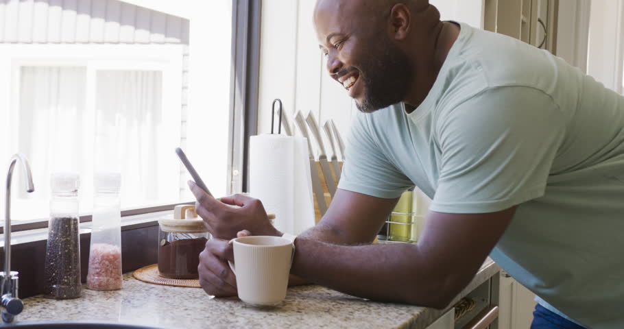 Smartphone buzzing African American midadult man in tee leaning on kitchen counter with mug reading. Phone, coffee, window, daylight, casual, lifestyle, cozy