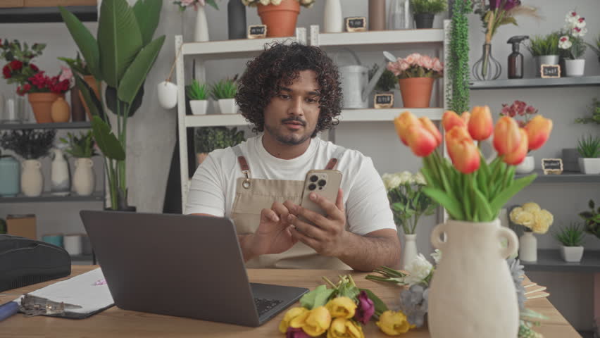 Man in apron holds smartphone and writes with pen while using laptop in a shop building arranging flowers on the counter; focused small business.