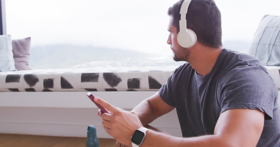 Adult man sitting right by window with headphones, glancing out, tapping phone, grabbing sunglasses. Smartphone, smartwatch, shades, contemplative, cozy, monochrome, nook