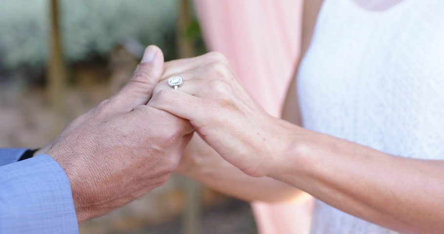 Man placing ring after vow exchange, woman smiling, couple sealing commitment beneath pink drapery. Outdoor, garden, ceremony, engagement, romance, dress, sunlight