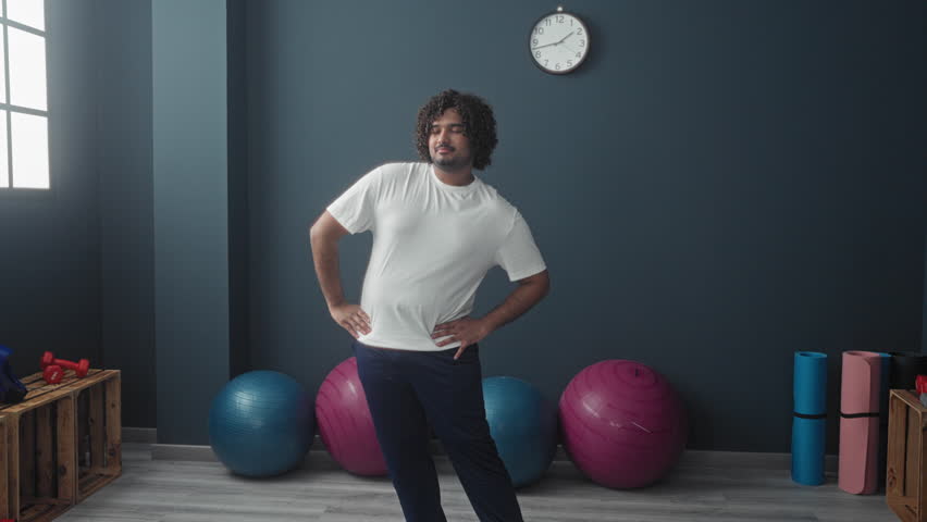 Young indian man standing with hands on hips by exercise and stability balls near window and clock in studio; confidence focus.