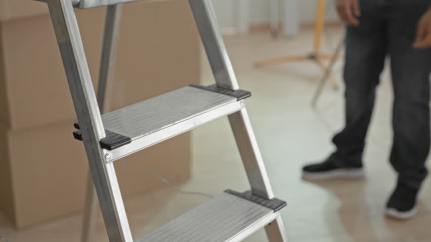 Young indian man standing with arms crossed beside stacked moving boxes inside a new home building, smiling with closed eyes as he surveys packed rooms; contentment.