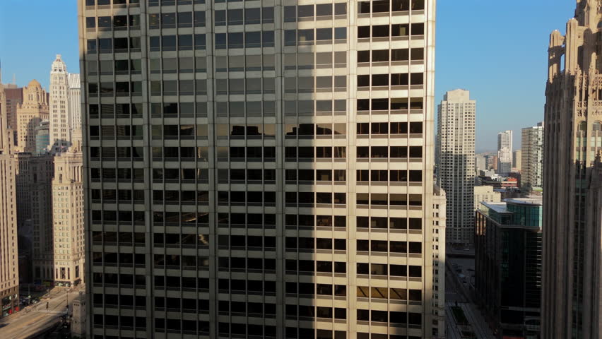 Drone footage reveals sleek glass facade of a modern office building in downtown Chicago. The reflective windows mirror surrounding historic architecture and Chicago River below on a clear sunny day.