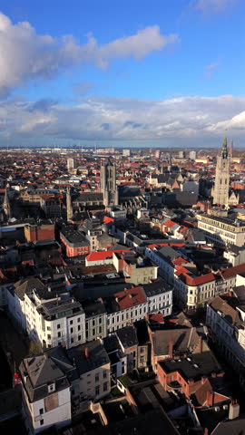 Vertical Screen: Aerial view of Ghent, Belgium showcasing historic gothic church towers, dense urban architecture with red tile rooftops, and dramatic cloudy sky over the medieval cityscape