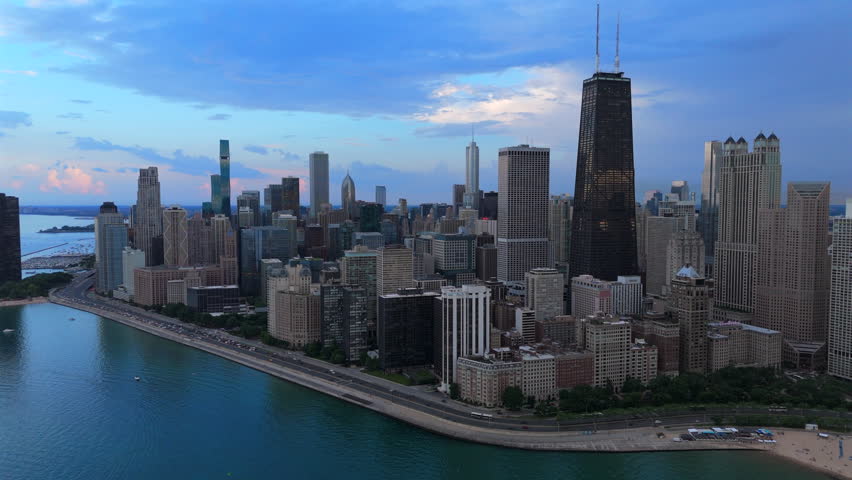 Stunning aerial view of Chicago downtown skyline along Lake Michigan shoreline at dusk. Tall skyscrapers dominate the cityscape with dramatic blue clouds overhead. Traffic flows on Lake Shore Drive.