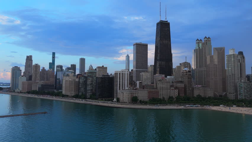 Stunning aerial view of Chicago skyline along Lake Michigan shoreline at dusk. Tall skyscrapers and iconic towers rise against dramatic blue cloudy sky. Urban waterfront with beach and highway.