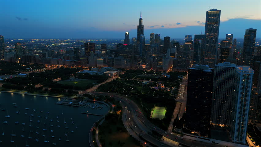 Stunning aerial view of Chicago skyline at twilight featuring illuminated skyscrapers, Grant Park, Lake Michigan harbor with boats, and city streets with traffic lights glowing at blue hour.