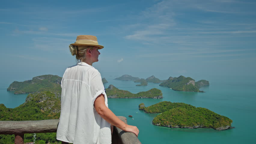 Middle-aged woman in a straw hat standing at a scenic viewpoint, looking over turquoise sea and tropical islands of Ang Thong Marine Park in Thailand.