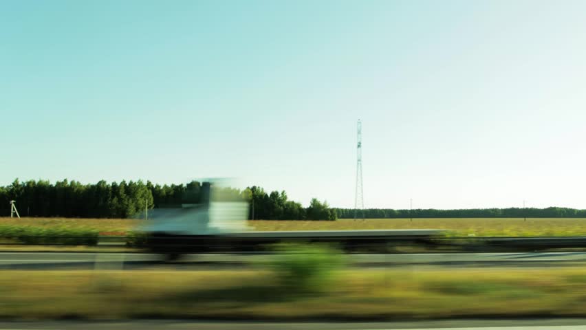 Dynamic side view from moving car. Roadside blur conveys speed. Summer landscape features vibrant green forest, large golden crop fields under bright sun, tall power transmission tower.