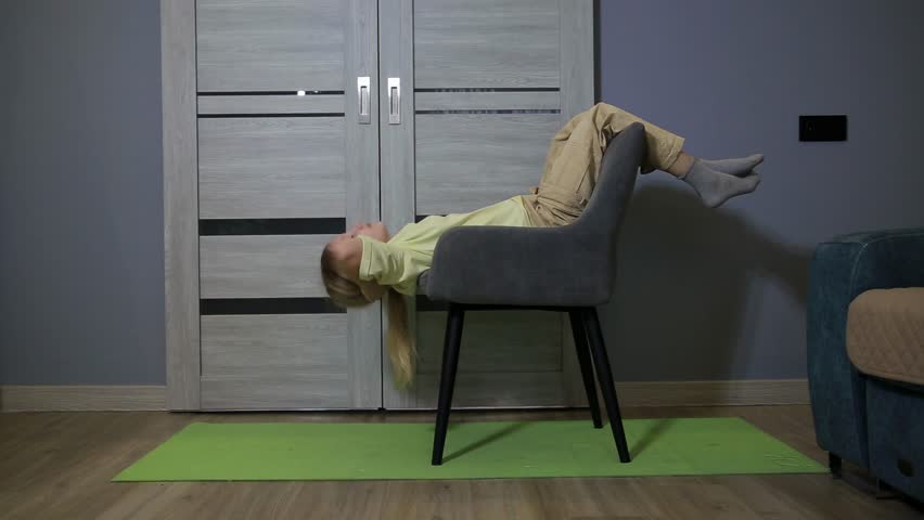 Full shot of young girl performing sit-ups using chair as support. Blonde hair child lying on chair with feet elevated for core strength training. Green mat on floor for exercise. Indoor fitness sessi