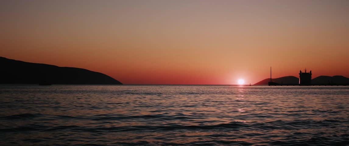 Sunset over sea with mountains and ship silhouette on horizon. Cinematic widescreen timelapse shot with anamorphic lens, orange sky reflection on water and calm seascape.
