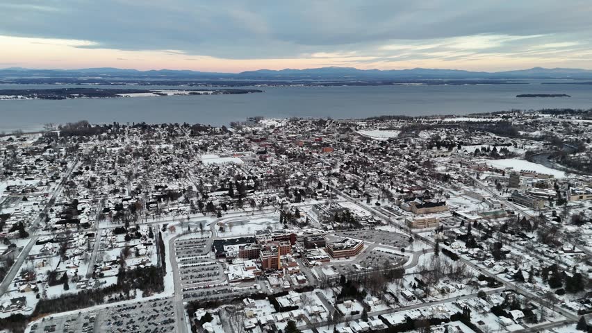 Dramatic aerial view showing the snow covered city of plattsburgh, new york, during a cold winter day with lake champlain and the adirondack mountains visible in the background