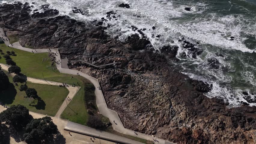 Aerial view of a rugged coastline meeting the foamy ocean, contrasted by green parkland and winding pathways filled with people, Porto, Portugal.