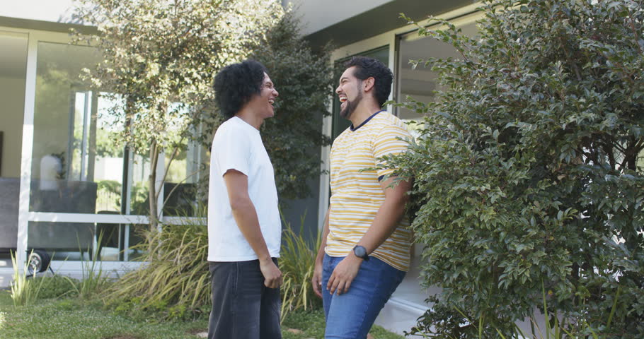 African American friends laughing after exchanging looks, stepping by window and shrub, posing. Duo, outdoor, yard, candid, casual, vibrant, modern