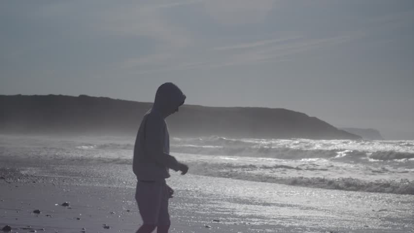 A silhouetted teenager in a hoodie walks along a beach, picking up and throwing stones into the rough, wavy ocean during the day.