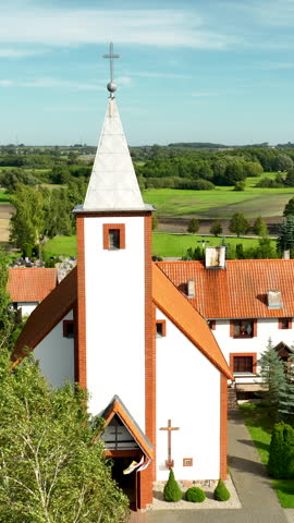 A vertical aerial shot of a charming white church with an orange tiled roof and a tall steeple, set against a lush green rural landscape in Poland.
