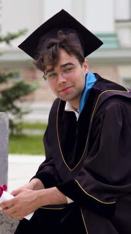 Male graduate in black cap and gown sits on a bench holding diploma with a proud expression, surrounded by greenery and modern architecture in the background