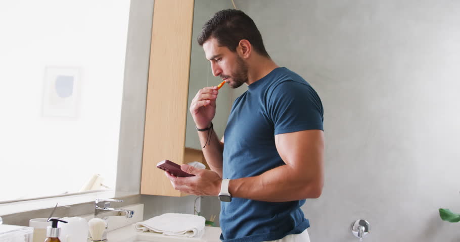 Adult man standing at sink by window, checking smartphone then brushing with orange toothbrush. Bathroom, phone, selfcare, grooming, routine, daylight, modern
