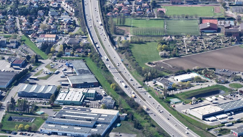 Aerial view of a highway cutting through the landscape, flanked by industrial buildings and green fields, creating a contrast, Sausheim, Grand Est, France.
