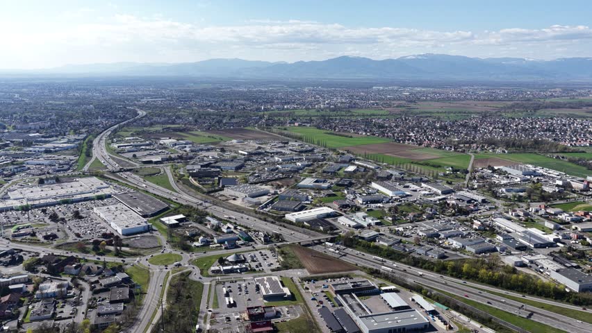 Aerial view of roads cutting through buildings with distant mountains under a blue sky, creating an interesting contrast, Sausheim, Grand Est, France.