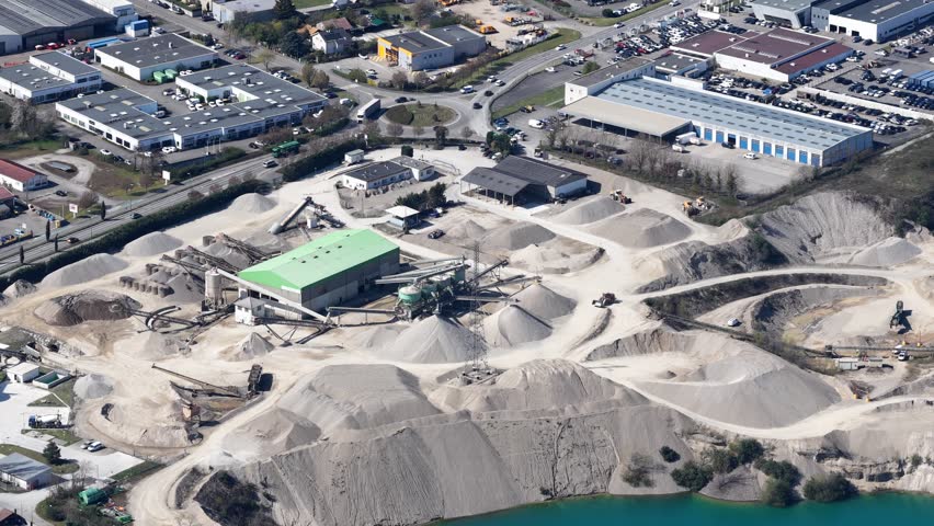 Aerial view of a vast quarry beside an industrial area, showcasing contrasting textures of gravel piles and buildings, Sausheim, Grand Est, France.