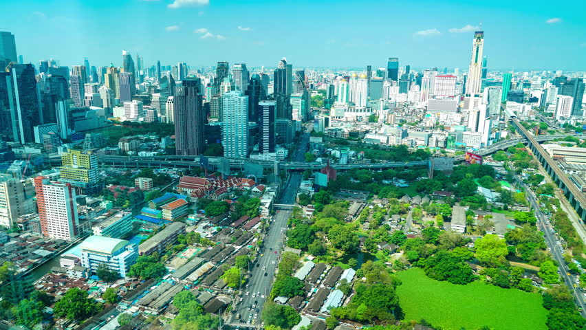 Aerial view of Bangkok skyline featuring modern skyscrapers, traditional temple complex, busy roads, and green urban spaces under bright clear sky